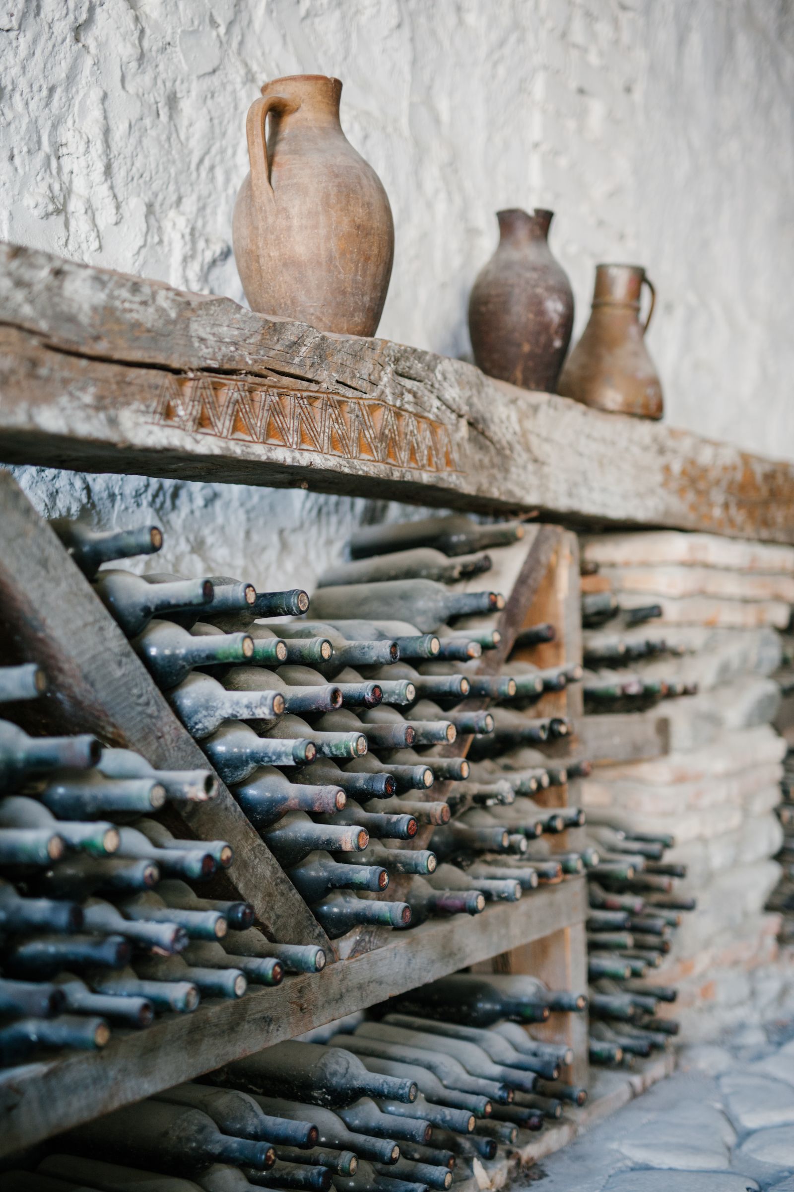 Botellas de vino antiguas cubiertas de polvo, tumbadas en un botellero de madera de un calado, con cántaros de barro sobre la repisa, imagen de las añadas guardadas de Rioja