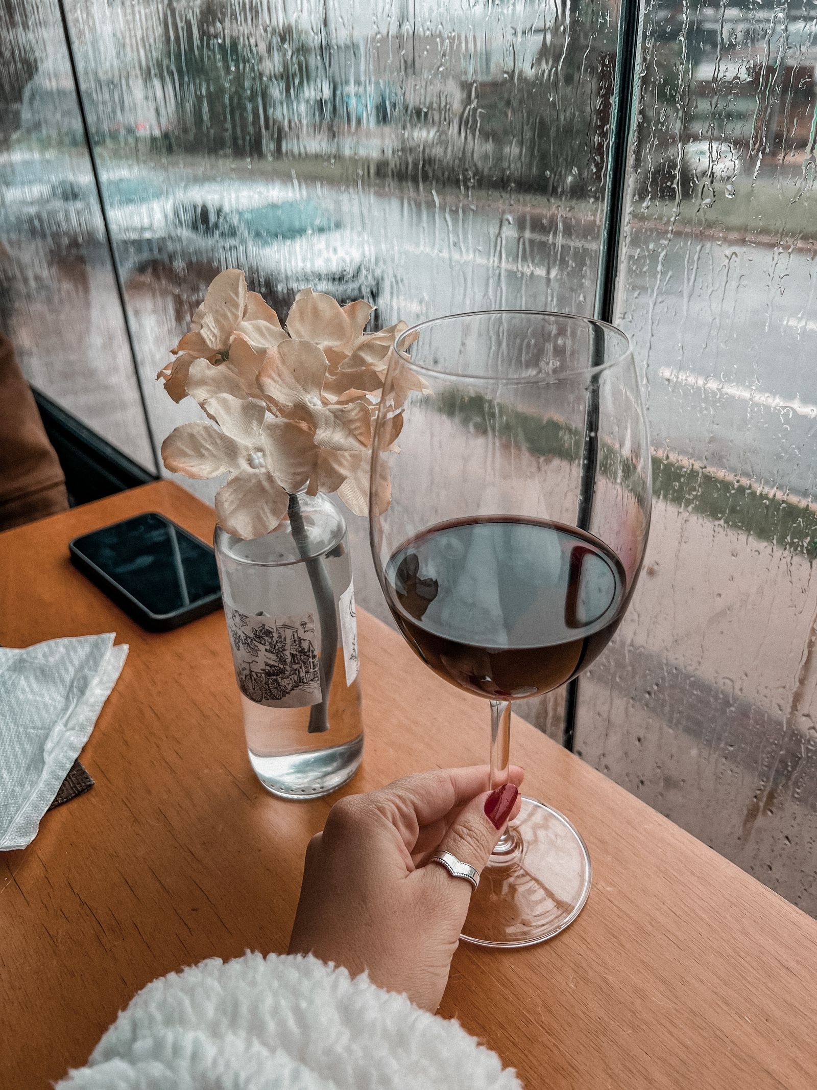 Copa de vino tinto sobre una mesa de madera junto a una ventana con gotas de lluvia en un día gris de otoño