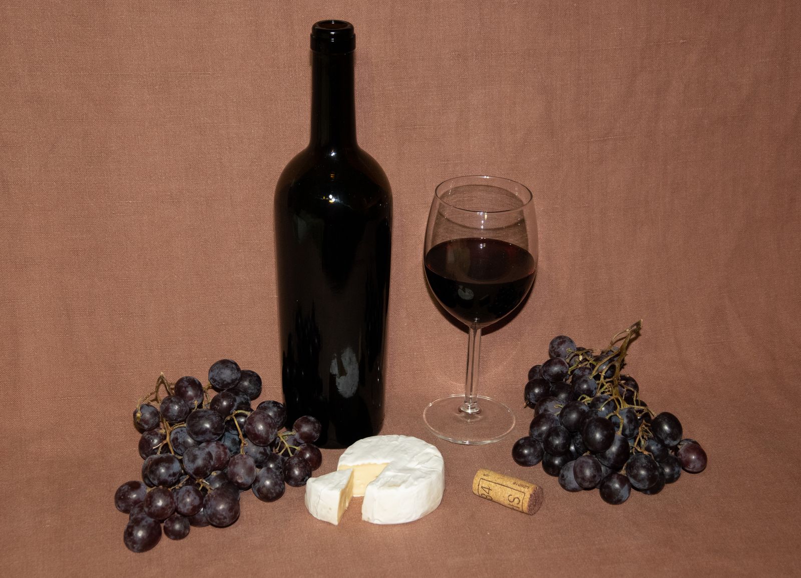 Botella de vino tinto sin etiqueta junto a una copa servida, un queso de corteza blanca cortado y racimos de uva tinta, preparados para una cata de queso y vino