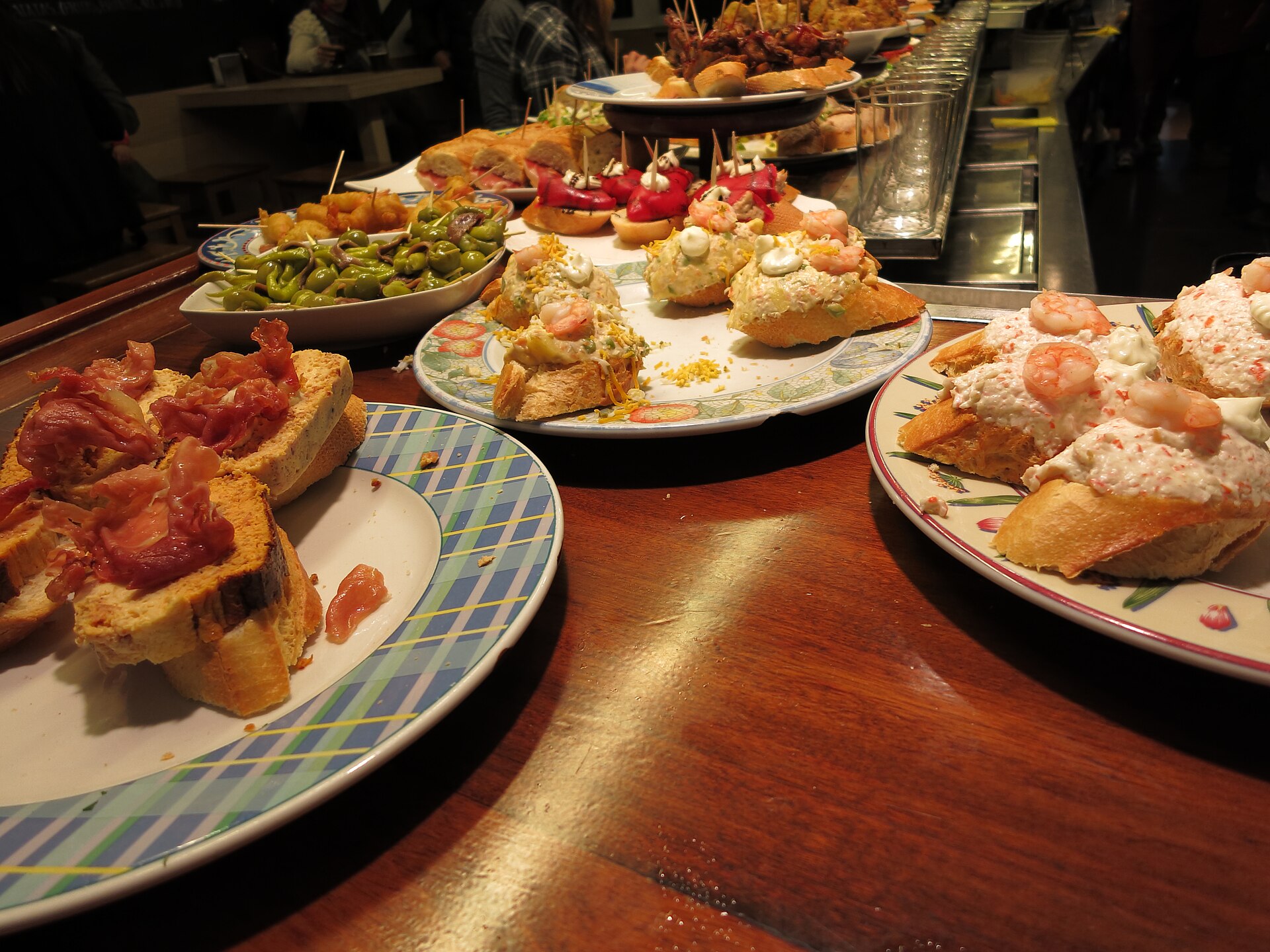 Barra de bar con platos de pinchos sobre pan, jamón, langostinos con salsa y guindillas, el tipo de barra que se recorre en una ruta de pinchos por La Herradura de Haro, La Rioja